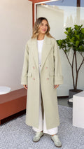 Front full view of woman wearing Sage Green double-breasted maxi trench coat with white wide-leg trousers